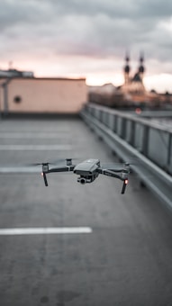 A drone hovers in focus above an empty rooftop parking lot with a blurred cityscape in the background under a cloudy sky.