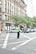 A city street scene with a traffic police officer standing in the middle of an intersection. The officer is wearing a reflective vest and directing traffic. Surrounding the intersection are several vehicles, including cars and a van. A large, ornate building with multiple windows is visible in the background, along with trees and additional urban structures.