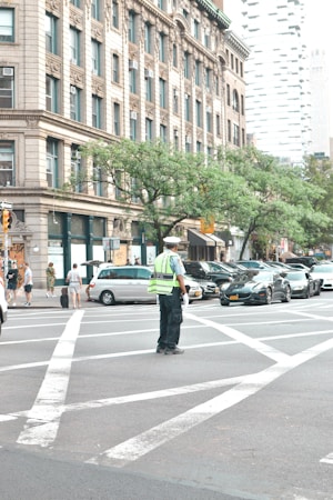 A city street scene with a traffic police officer standing in the middle of an intersection. The officer is wearing a reflective vest and directing traffic. Surrounding the intersection are several vehicles, including cars and a van. A large, ornate building with multiple windows is visible in the background, along with trees and additional urban structures.