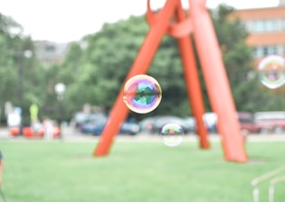 A large soap bubble floats in the foreground with a vibrant rainbow reflection on its surface. In the background, a red-orange metal sculpture is present against a backdrop of green trees and a blurred urban environment.