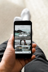 A person holding a smartphone displaying an Instagram profile with a grid of various outdoor and landscape photos. The person appears to be sitting with one leg crossed over the other, dressed in dark pants and white socks. The focus is on the phone, which is centered in the frame.