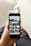 A person holding a smartphone displaying an Instagram profile with a grid of various outdoor and landscape photos. The person appears to be sitting with one leg crossed over the other, dressed in dark pants and white socks. The focus is on the phone, which is centered in the frame.