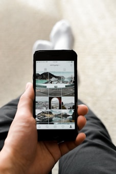A person holding a smartphone displaying an Instagram profile with a grid of various outdoor and landscape photos. The person appears to be sitting with one leg crossed over the other, dressed in dark pants and white socks. The focus is on the phone, which is centered in the frame.
