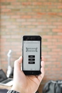 A person is holding a smartphone displaying an app with buttons labeled Scan, Cart, and History against a blurred brick wall background. The app interface appears to be minimalistic and functional.