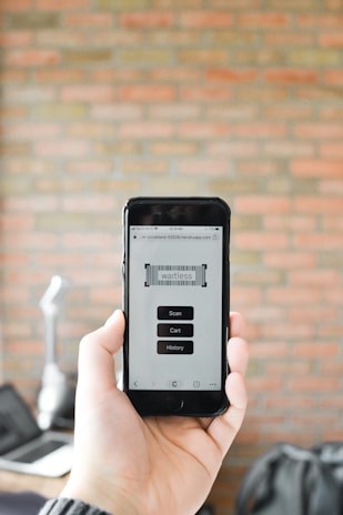 A person is holding a smartphone displaying an app with buttons labeled Scan, Cart, and History against a blurred brick wall background. The app interface appears to be minimalistic and functional.