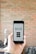 A person is holding a smartphone displaying an app with buttons labeled Scan, Cart, and History against a blurred brick wall background. The app interface appears to be minimalistic and functional.
