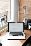 A laptop displaying a graph is placed on a wooden desk near a window. To the left, a shelf holds a small clock and a pen. A desk lamp and speaker are situated to the right. The wall in the background is made of light-colored bricks.