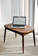 A laptop is placed on a small wooden table in a room with light-colored carpet and vertical blinds. The laptop screen displays a webpage titled 'Interactive RFID', indicating a focus on technology.