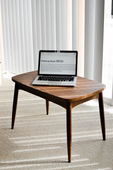 A laptop is placed on a small wooden table in a room with light-colored carpet and vertical blinds. The laptop screen displays a webpage titled 'Interactive RFID', indicating a focus on technology.