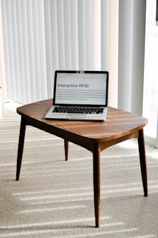 A laptop is placed on a small wooden table in a room with light-colored carpet and vertical blinds. The laptop screen displays a webpage titled 'Interactive RFID', indicating a focus on technology.