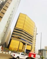 A modern office building with reflective golden-tinted glass panels dominates the scene. The structure is surrounded by other high-rise buildings, indicating an urban environment. In the foreground, a white Toyota pickup truck and a red Airtel-branded kiosk are visible on the road.