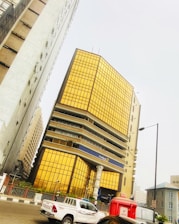 A modern office building with reflective golden-tinted glass panels dominates the scene. The structure is surrounded by other high-rise buildings, indicating an urban environment. In the foreground, a white Toyota pickup truck and a red Airtel-branded kiosk are visible on the road.