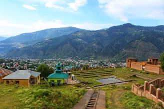 A scenic landscape featuring a mountainous backdrop with a lush green valley. In the foreground, there are historic ruins with brick structures and a geometric garden. Some residential houses with colorful roofs are visible, surrounded by trees.