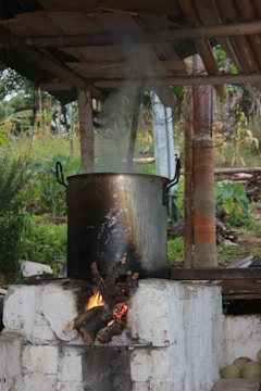 A large metal pot sits on a brick stove with a wood fire burning underneath. Smoke rises from the pot, indicating something is being cooked. The setup is outdoors under a simple wooden shelter, surrounded by greenery and some rural vegetation.