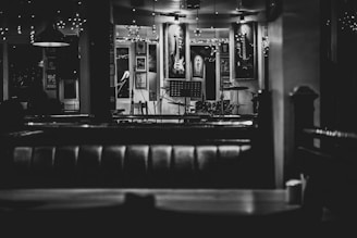 An empty, dimly lit bar or cafe with a stage set up for live music. Musical instruments, including a guitar on the wall and a drum set on stage, are visible. The decor includes framed pictures and string lights, creating a cozy and intimate atmosphere.