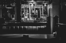 An empty, dimly lit bar or cafe with a stage set up for live music. Musical instruments, including a guitar on the wall and a drum set on stage, are visible. The decor includes framed pictures and string lights, creating a cozy and intimate atmosphere.