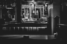 An empty, dimly lit bar or cafe with a stage set up for live music. Musical instruments, including a guitar on the wall and a drum set on stage, are visible. The decor includes framed pictures and string lights, creating a cozy and intimate atmosphere.