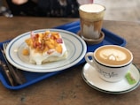 A wooden table with a blue tray holding a plate of dessert and two beverages. The dessert appears to be a fluffy cake topped with cream and diced peaches. One beverage is a cappuccino with latte art served in a white cup with a saucer, accompanied by a sugar packet. The other drink appears to be a frothy coffee served in a glass.