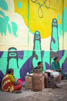 Three women are sitting on the ground near a colorful mural. One woman is wearing a red and green sari and is holding a cell phone. The other two women are seated next to a makeshift cooking area with metal pots, a gas cylinder, and wooden crates. The mural behind them is bright with abstract shapes and bold colors, featuring large bottle-like forms in teal and black.