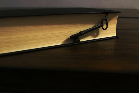 A thick, hardcover book lying closed on a wooden table with an old-fashioned skeleton key placed on its surface. The book's pages are a light cream color, contrasting with its dark cover. The key casts a shadow, indicating a warm, soft light source.