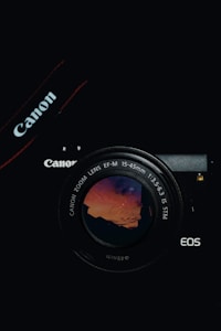 A Canon camera with a lens reflecting a vivid sunset sky is positioned against a dark backdrop. The camera's branding is visible on the strap and body.