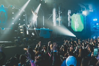 A lively concert scene with a large crowd of people raising their hands and smartphones, capturing the moment on stage. A performer stands on an elevated platform, reaching out energetically.