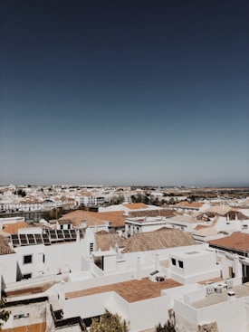 A panoramic view of a coastal town featuring white buildings with terra-cotta tiled roofs. The scene includes solar panels on some rooftops and a distant river or waterway. The sky is clear and blue, adding to the serene atmosphere.