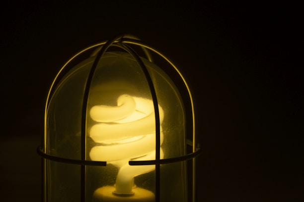 A spiral fluorescent light bulb is encased within a protective wire guard, emitting a soft yellow glow against a dark background. The light is partially visible through the wire, emphasizing the warm illumination.