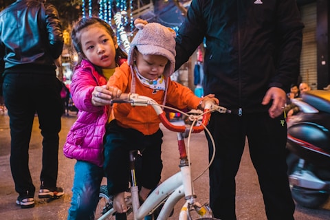 Two children are positioned on a bicycle, with one child sitting on the seat and the other standing behind, holding onto the handlebars. A person in a black tracksuit stands nearby, providing support. The scene is set on a city street, illuminated by festive string lights in the background with several pedestrians walking by.