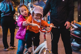 Two children are positioned on a bicycle, with one child sitting on the seat and the other standing behind, holding onto the handlebars. A person in a black tracksuit stands nearby, providing support. The scene is set on a city street, illuminated by festive string lights in the background with several pedestrians walking by.