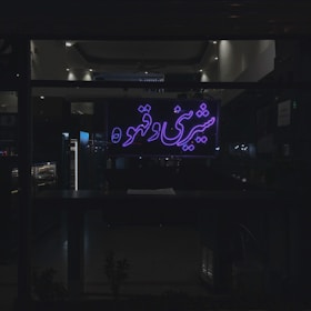 A dimly lit interior space is visible through a large window, which features a neon sign with Persian script glowing in purple. The environment inside appears to be a café or shop, with shelves and lighting fixtures indistinctly visible in the background.