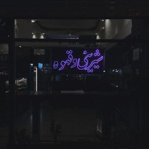 A dimly lit interior space is visible through a large window, which features a neon sign with Persian script glowing in purple. The environment inside appears to be a café or shop, with shelves and lighting fixtures indistinctly visible in the background.