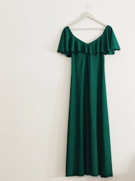 A long, green off-the-shoulder dress with ruffle details hangs on a white hanger against a light-colored wall.