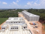 Aerial view of a construction site with several rectangular buildings in various stages of completion. The structures are surrounded by a mix of cleared land and dense green forest. A dirt road runs through the middle, with construction materials and cargo containers visible along the sides.