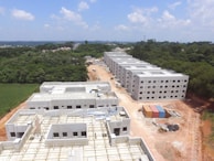 Aerial view of a construction site with several rectangular buildings in various stages of completion. The structures are surrounded by a mix of cleared land and dense green forest. A dirt road runs through the middle, with construction materials and cargo containers visible along the sides.