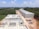 Aerial view of a construction site with several rectangular buildings in various stages of completion. The structures are surrounded by a mix of cleared land and dense green forest. A dirt road runs through the middle, with construction materials and cargo containers visible along the sides.