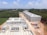 Aerial view of a construction site with several rectangular buildings in various stages of completion. The structures are surrounded by a mix of cleared land and dense green forest. A dirt road runs through the middle, with construction materials and cargo containers visible along the sides.