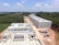 Aerial view of a construction site with several rectangular buildings in various stages of completion. The structures are surrounded by a mix of cleared land and dense green forest. A dirt road runs through the middle, with construction materials and cargo containers visible along the sides.
