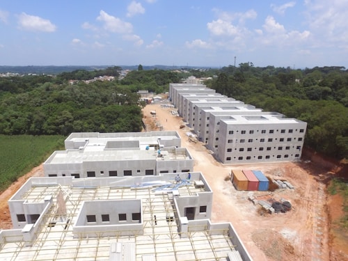 Aerial view of a construction site with several rectangular buildings in various stages of completion. The structures are surrounded by a mix of cleared land and dense green forest. A dirt road runs through the middle, with construction materials and cargo containers visible along the sides.