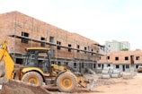 A construction site with a partially built brick building featuring metal scaffolding on the exterior. A large yellow backhoe loader is parked in front of the building. Several concrete pipes are stacked nearby along with other construction materials scattered around the unfinished structure.