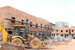 A construction site with a partially built brick building featuring metal scaffolding on the exterior. A large yellow backhoe loader is parked in front of the building. Several concrete pipes are stacked nearby along with other construction materials scattered around the unfinished structure.