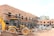 A construction site with a partially built brick building featuring metal scaffolding on the exterior. A large yellow backhoe loader is parked in front of the building. Several concrete pipes are stacked nearby along with other construction materials scattered around the unfinished structure.