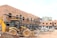 A construction site with a partially built brick building featuring metal scaffolding on the exterior. A large yellow backhoe loader is parked in front of the building. Several concrete pipes are stacked nearby along with other construction materials scattered around the unfinished structure.