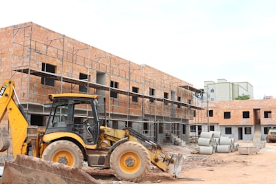 A construction site with a partially built brick building featuring metal scaffolding on the exterior. A large yellow backhoe loader is parked in front of the building. Several concrete pipes are stacked nearby along with other construction materials scattered around the unfinished structure.