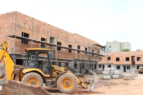 A construction site with a partially built brick building featuring metal scaffolding on the exterior. A large yellow backhoe loader is parked in front of the building. Several concrete pipes are stacked nearby along with other construction materials scattered around the unfinished structure.