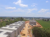 A construction site with several unfinished buildings arranged in rows is visible. The buildings are surrounded by a mixture of green areas, trees, and farmland extending into the horizon. A dirt road runs through the center of the site, with construction equipment visible, including a yellow bulldozer. The sky is mostly clear with some scattered clouds.