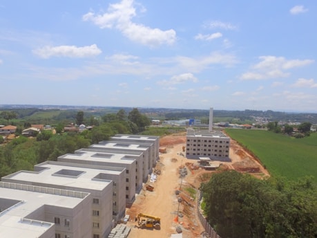 A construction site with several unfinished buildings arranged in rows is visible. The buildings are surrounded by a mixture of green areas, trees, and farmland extending into the horizon. A dirt road runs through the center of the site, with construction equipment visible, including a yellow bulldozer. The sky is mostly clear with some scattered clouds.