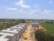 A construction site with several unfinished buildings arranged in rows is visible. The buildings are surrounded by a mixture of green areas, trees, and farmland extending into the horizon. A dirt road runs through the center of the site, with construction equipment visible, including a yellow bulldozer. The sky is mostly clear with some scattered clouds.