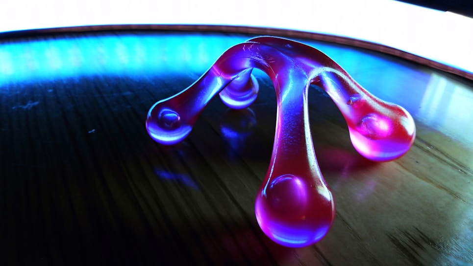 A translucent, purple massager with four rounded ends rests on a wooden surface. The background is illuminated with a bright blue and white light, creating a striking reflective effect on the surface.