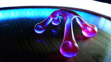 A translucent, purple massager with four rounded ends rests on a wooden surface. The background is illuminated with a bright blue and white light, creating a striking reflective effect on the surface.